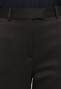 Detailný záber na tmavohnedé nohavice s jemnými vertikálnymi prúžkami a pásom s pútkami na opasok, kombinované s tmavým pleteným topom.