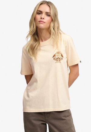 Lyshåret kvinde med løst hår iført en beige t-shirt med en brun emblemlogo på venstre bryst og brune bukser, stående med hænderne bag ryggen.