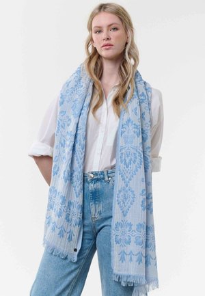 Young woman with long blonde hair wearing a white button-up shirt, blue jeans, and a light blue patterned scarf draped over her shoulders.