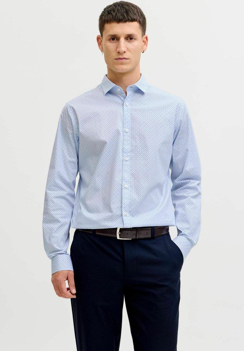 Mlad moški stoji, oblečen v svetlo modro s pikami obarvano srajco, zatlačeno v črne hlače s črnim pasom, roke sproščene ob straneh.