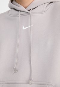 Grå Nike-hoodie med en vit swoosh-logga, snörningar med knutade ändar och ett mjukt, texturerat tyg.