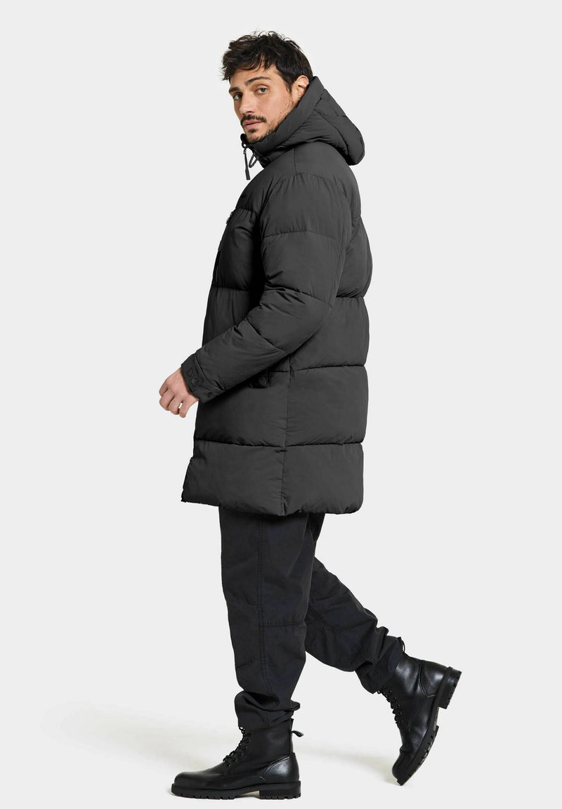 Schwarze Daunenjacke mit Kapuze, ausgestattet mit einem gesteppten Design und einer leicht verlängerten Silhouette. Kombiniert mit schwarzen Hosen und Stiefeln.