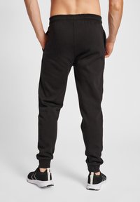 Schwarze Baumwoll-Sweatpants mit geripptem Bund, seitlichen Taschen und tailliertem Schnitt. Kombiniert mit schwarzen Sportschuhen mit weißen Akzenten.