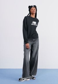 Fekete kapucnis felső fehér "new balance" logóval, szürke, bő szárú farmernadrág és fekete-fehér sportcipő. A modell fekete sapkát visel.