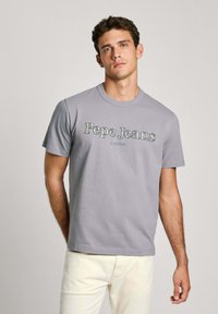 Šedé bavlněné tričko s potiskem "Pepe Jeans" a "London" v kontrastních písmenech. Umístěné na neutrálním pozadí. Krátké rukávy.