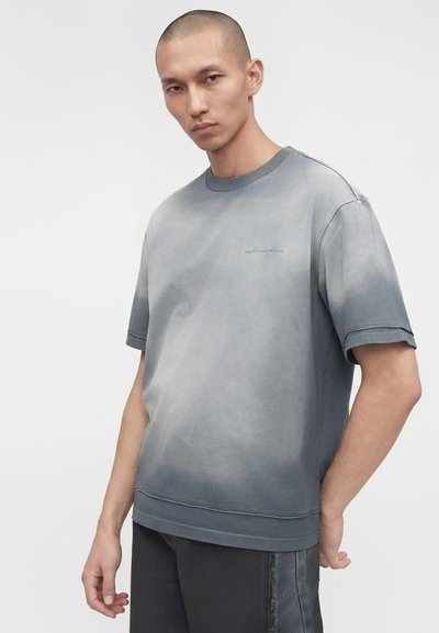 Άνδρας που φοράει ξεθωριασμένο γκρι κοντομάνικο μπλουζάκι και σκούρο παντελόνι, στέκεται με το ένα χέρι πίσω από την πλάτη, ουδέτερη έκφραση, ανοιχτό γκρι φόντο.