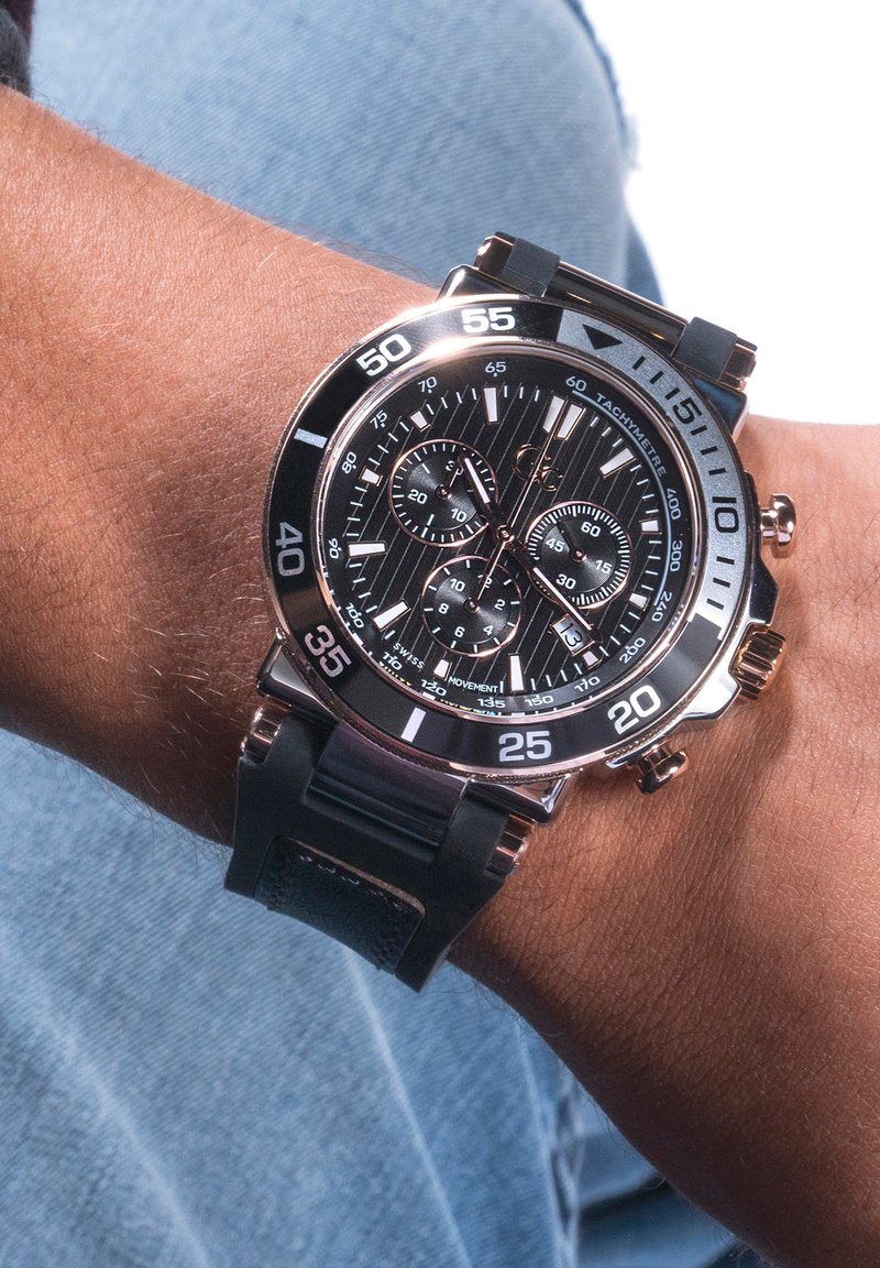 Zwarte chronograafhorloge met een gestructureerde wijzerplaat, rosegouden accenten, tachymeterschaal en een zwarte rubberen band, weergegeven op een pols.