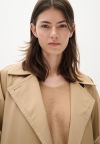 Beige trench coat with wide lapels, paired with a light brown knit sweater. Smooth fabric texture and minimalistic design.