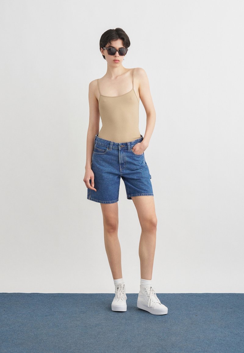 Personne aux cheveux courts et foncés portant des lunettes de soleil noires, un débardeur beige, un short en jean bleu, des chaussettes blanches et des baskets blanches, debout sur un tapis bleu.