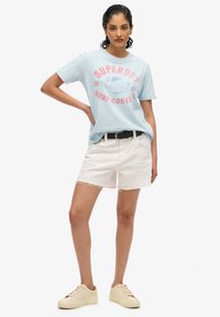 Camiseta de algodón azul claro con estampado gráfico rosa, combinada con pantalones cortos de mezclilla blancos. La modelo lleva un cinturón negro y zapatillas beige.