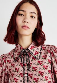 Blusa floreale in tessuto rosa con maniche a sbuffo, dettagli neri e un colletto. Caratterizzata da fiori rossi e foglie verdi in un motivo a griglia.