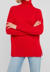 Pull en tricot rouge à col montant, texture côtelée et coutures d'épaules tombantes. Présente un ourlet côtelé et des fentes sur les côtés pour plus de confort.