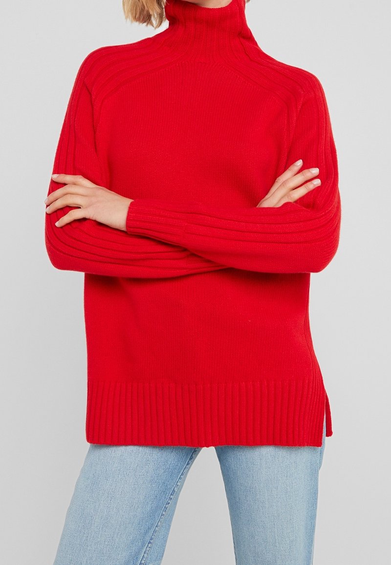 Pull en tricot rouge à col montant, texture côtelée et coutures d'épaules tombantes. Présente un ourlet côtelé et des fentes sur les côtés pour plus de confort.