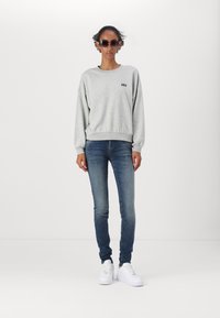 Szürke pamut pulóver GAP logóval, kék skinny farmernadrággal és fehér sneakerrel párosítva. Modell fekete napszemüveget visel, egy egyszínű háttér előtt áll.