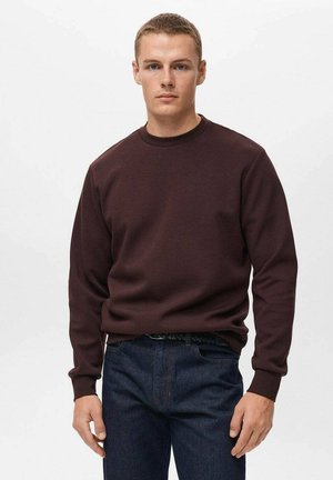 Suéter burdeos con cuello redondo y mangas largas, hecho de un material suave. Llevado con jeans azul oscuro, que cuentan con un cinturón texturizado.