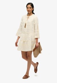 Hellbeige gestufte Kleid mit Bindegürtel und Quasten-Details, ausgestattet mit Glockenärmeln und dezenten strukturierten Mustern. Die Accessoires bestehen aus einer geflochtenen Clutch und Sandalen.