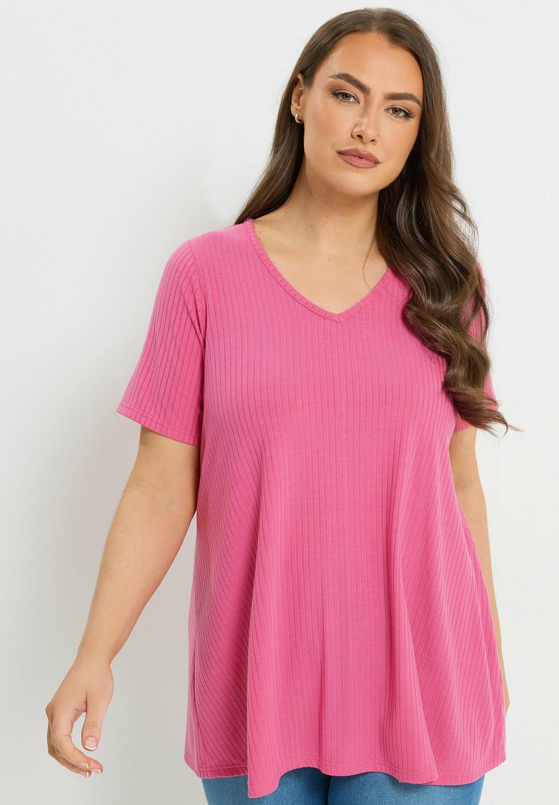 Frau mit langem, welligem Haar, die ein geripptes, kurzärmliges, rosa V-Ausschnitt-Oberteil und blaue Jeans trägt, steht vor einem schlichten hellen Hintergrund.