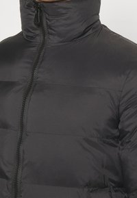 Svart vadderad jacka med hög krage och central dragkedja, bärs av en person som visar övre delen av bröstet och nackområdet.