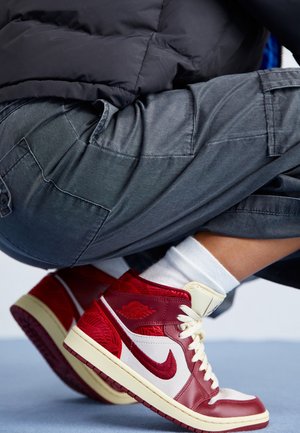 Persoon die hurkt en rode en witte Nike Air Jordan sneakers, witte sokken, grijze cargobroek en een donker jack draagt.