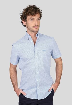 Chemise bleu clair à manches courtes avec un motif à pois subtil, col boutonné et une petite poche sur la poitrine gauche. Texture de tissu lisse.