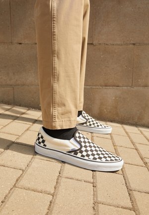 Baskets à enfiler en damier noir et blanc avec une semelle en caoutchouc blanc, portées avec un pantalon kaki et des chaussettes noires, se tenant sur une surface pavée.