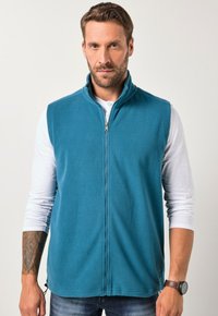 Fleece-Weste in Teal mit Stehkragen und einem Frontreißverschluss. Sie hat eine glatte Textur und ist über einem weißen Langarmshirt getragen.