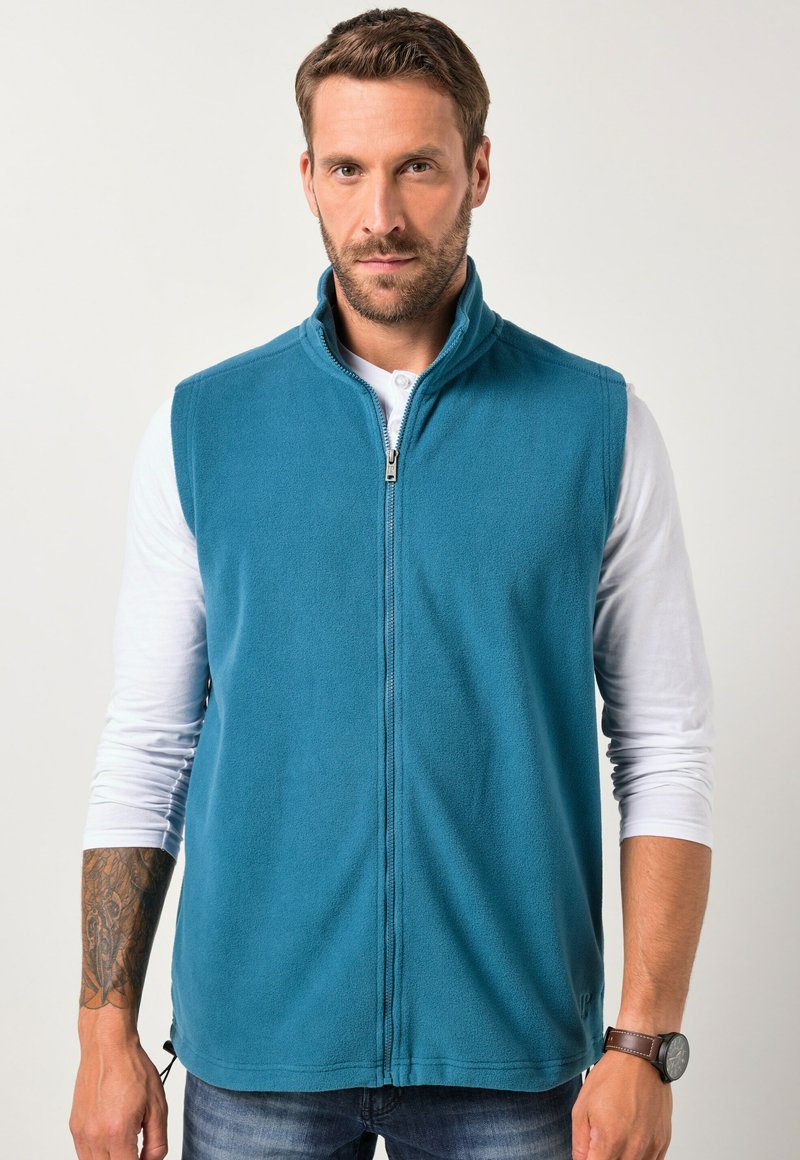 Fleece-Weste in Teal mit Stehkragen und einem Frontreißverschluss. Sie hat eine glatte Textur und ist über einem weißen Langarmshirt getragen.