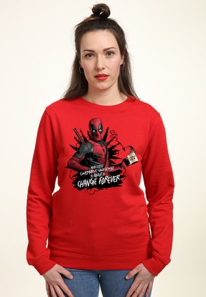Kvinde med mørkt hår i en topknude har en rød Deadpool-sweatshirt med teksten "Dit lille filmiske univers er ved at ændre sig for evigt."