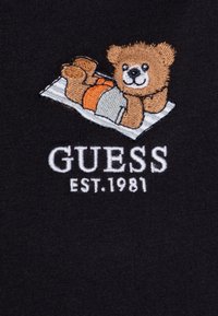 Iegūts brūnā teddy lāča izšuvums uz melna auduma fona, ar oranžiem un pelēkiem akcentiem, un tekstu "GUESS EST. 1981" zem tā.