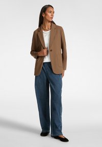 Blazer marrone con un solo bottone, abbinato a un top bianco e pantaloni in denim a gamba larga con un dettaglio di perle lungo il lato. Ballerine nere.