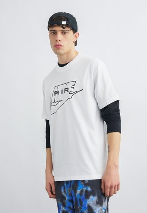 Fiatal férfi fekete sapkát visel hátrafelé, fehér Nike Air pólót fekete hosszú ujjú ing fölött, és kék mintás nadrágot, egyszínű háttér előtt.