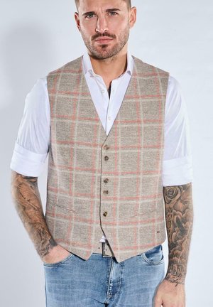 Man with rolled-up white shirt sleeves wears beige plaid vest and blue jeans, showing detailed sleeve tattoos on both arms.