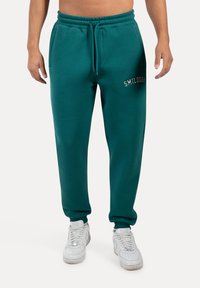 Joggers de algodón en color azul verdoso con cinturilla elástica, bolsillos laterales y "SMILDOOX" bordado en blanco en el muslo superior. Combinados con zapatillas blancas.