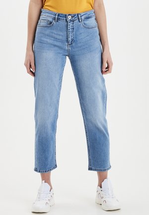 Persoon die lichtblauwe, hoogwater, cropped jeans, een geel shirt en witte sneakers draagt, staand op een effen witte achtergrond.