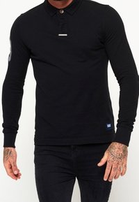 Polo noir à manches longues avec col à boutons, présentant un accent en métal, des poignets côtelés et une petite étiquette logo à l'ourlet.
