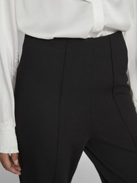 Black high-waisted trousers with clean lines, made of textured fabric. Paired with a white, silky blouse featuring gathered cuffs.