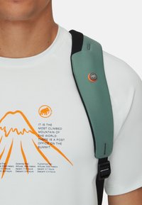 Teal axelrem fäst vid en svart ryggsäck, med en orange cirkulär detalj. Personen bär en vit t-shirt med ett bergsgrafik och text.