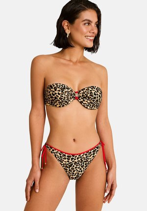 Femme souriante, portant un bikini bandeau à imprimé léopard avec des bordures rouges et des liens latéraux sur le bas, debout devant un fond uni.