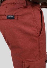 Pantalons couleur rouille en tissu résistant, dotés d'une poche latérale, d'une étiquette tissée au dos et d'une finition texturée.