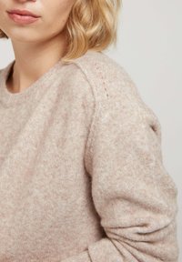 Un pull en tricot rose clair avec une finition texturée, un col rond et des coutures raglan subtiles. Le tissu a l'air doux et confortable.