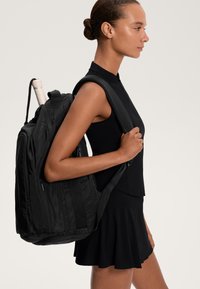Fekete nejlon hátizsák párnázott pántokkal, elegáns dizájnnal és külső cipzáras zsebekkel. Egy vízpalack részben látható az oldalsó zsebben.