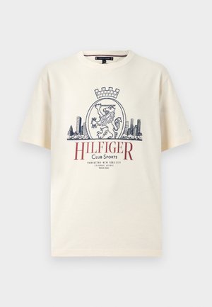 Cremefarbenes Baumwoll-T-Shirt mit kurzen Ärmeln, mit einer marineblauen Grafik eines Löwen, einer Krone und einer Stadtsilhouette, darunter roter Text.