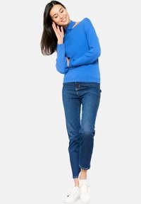 Pull bleu à col montant, manches longues et poignets côtelés, associé à un jean bleu et des baskets blanches.