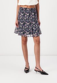 Jupe à motif floral en bleu foncé, en tissu léger, avec une taille froncée, de forme A-line fluide, assortie à des escarpins noirs à bout pointu.