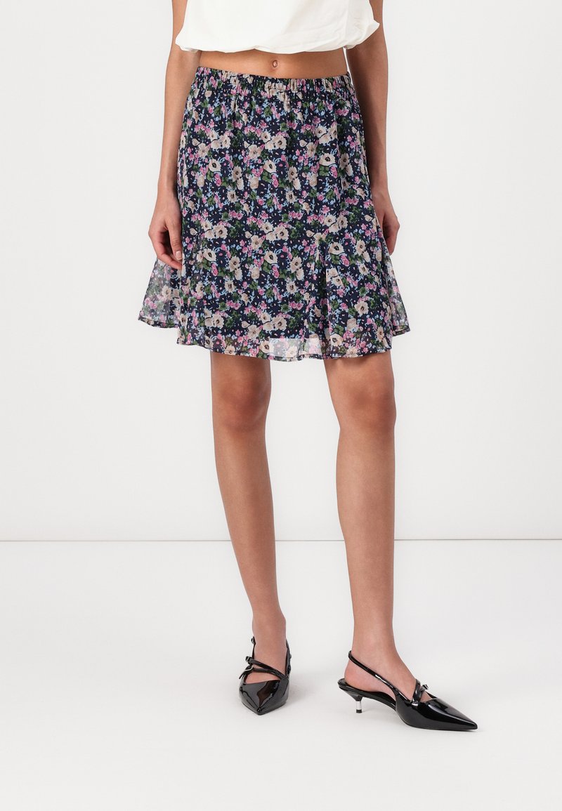 Jupe à motif floral en bleu foncé, en tissu léger, avec une taille froncée, de forme A-line fluide, assortie à des escarpins noirs à bout pointu.