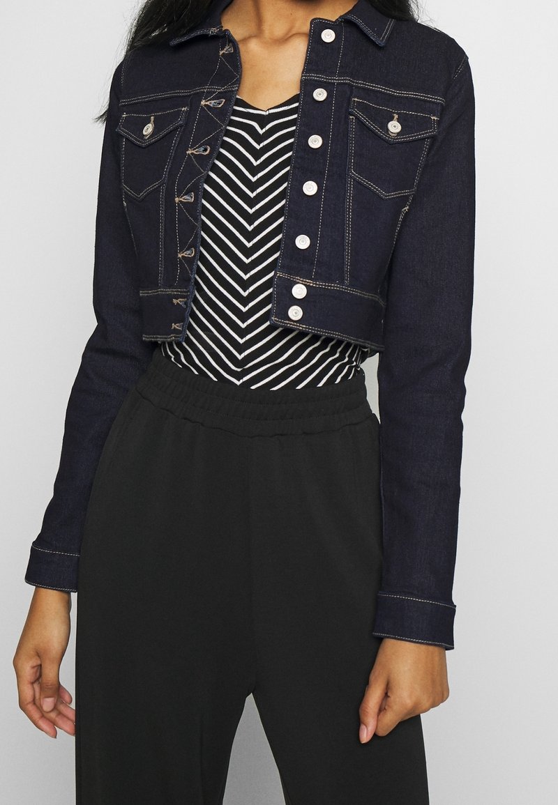 Femme portant une veste courte en denim foncé sur un haut noir et blanc à motif chevron avec un pantalon noir taille haute.
