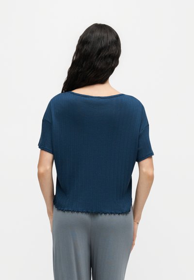 Femme aux longs cheveux foncés portant un haut bleu ample à manches courtes avec un ourlet légèrement froncé et un pantalon gris clair, vue de dos sur fond blanc.