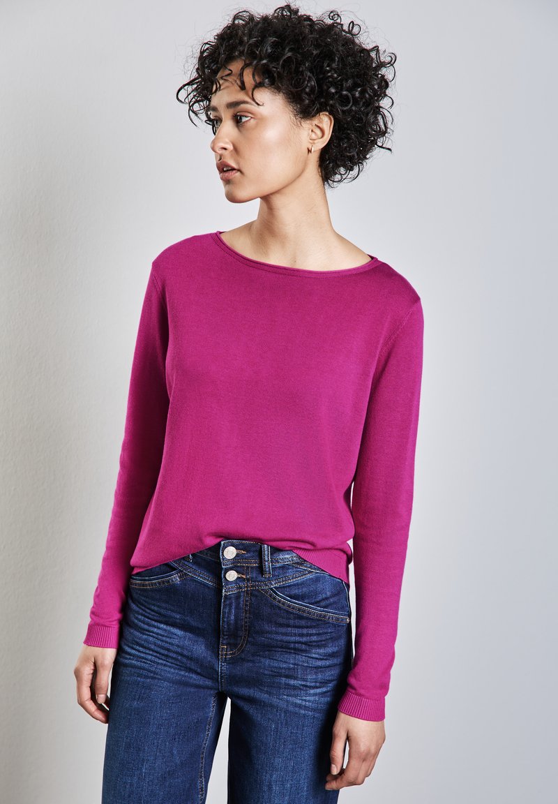 Donna con capelli corti e ricci che indossa una maglietta a maniche lunghe rosa acceso e jeans blu scuro, che guarda verso sinistra su uno sfondo semplice.