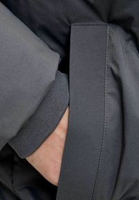 Szara kurtka o gładkim, matowym wykończeniu. Posiada ściągacz przy mankietach oraz boczną kieszeń, z widocznymi czystymi szwami i nowoczesnym designem.
