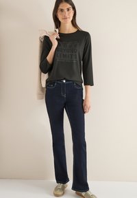 Schwarzes Dreiviertelarm-Oberteil mit grafischem Text kombiniert mit dunkelblauen Jeans. Das Modell trägt hellfarbene Turnschuhe und hält einen beigen Pullover in der Hand.